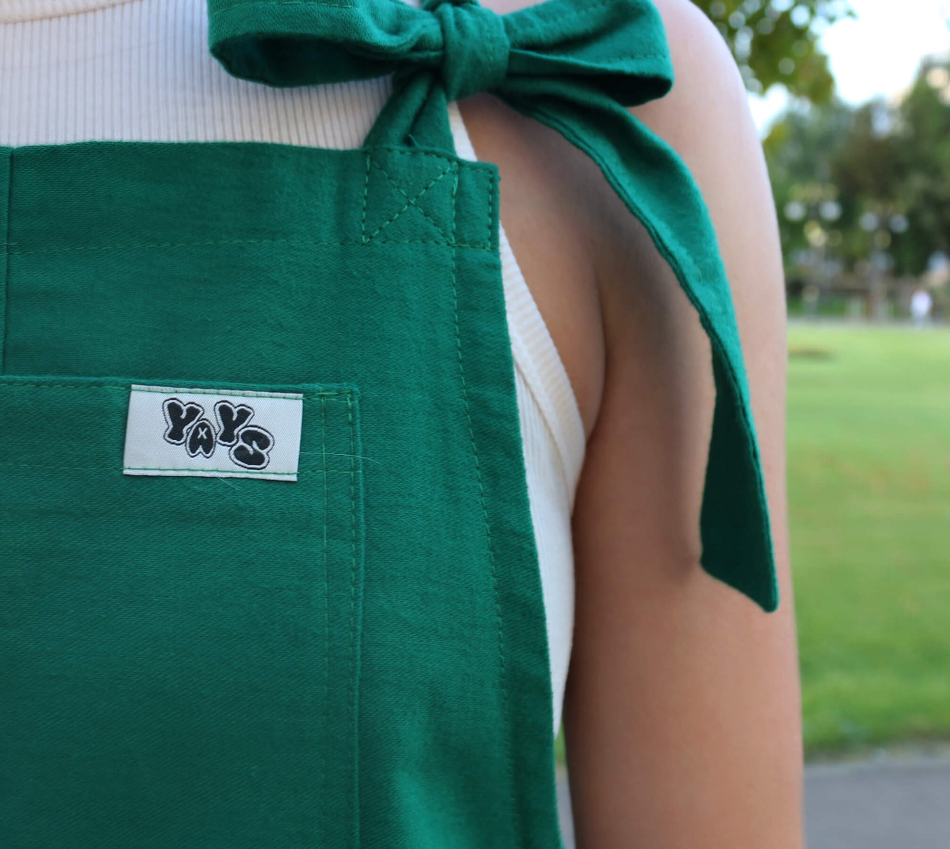 woman wearing YAYS linen unisex dungarees in Spring Green, posing extra close-up showing details of front pocket, bow on adjustable straps and YAYS logo